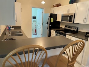 A kitchen with white cabinets, a stainless steel sink, a microwave, and a stove. There are two wooden chairs and a small island with a countertop.