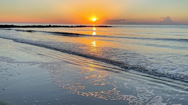 A beach with a sunset over the ocean, waves gently lapping at the shore, and a clear sky.