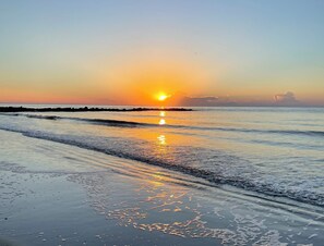 Ein Strand mit Sonnenuntergang über dem Ozean, sanfte Wellen schlappen ans Ufer und der Himmel ist klar.