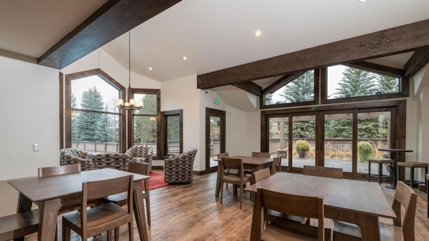 Spaziosa sala da pranzo con tavoli e sedie in legno, ampie finestre con vista sugli alberi e un soffitto alto con travi a vista.