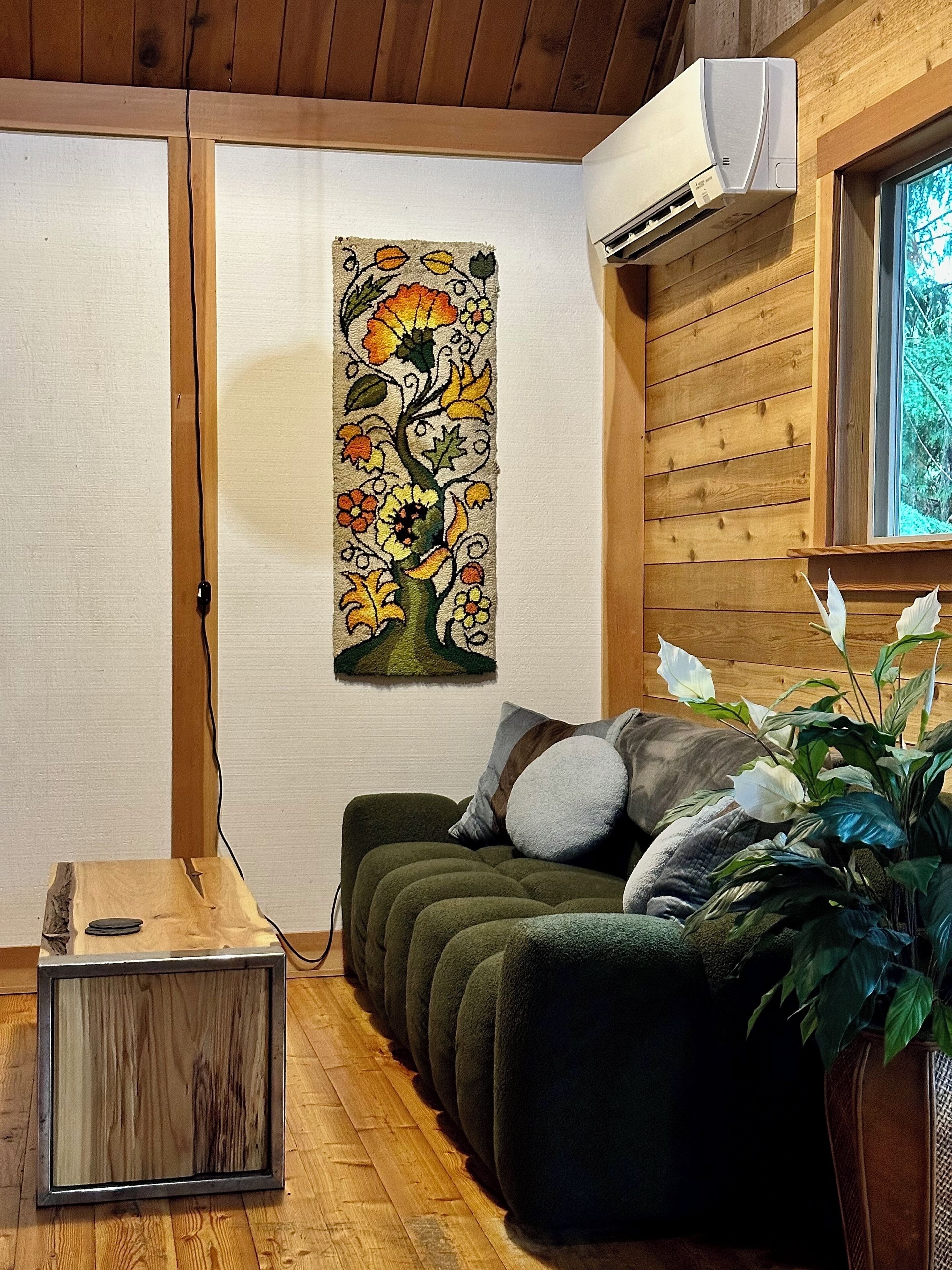 A cozy room with a wooden ceiling, a wall-mounted air conditioner, a green sofa with cushions, a framed floral tapestry, and a wooden cabinet with a mirror.