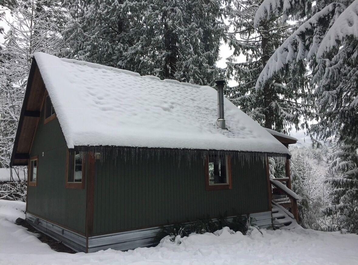 A small, single-story cabin with a steeply pitched roof, surrounded by snow-covered trees.