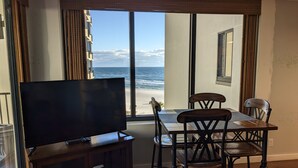 A room with a TV on a wooden stand, a dining table with chairs, and a view of the beach through large windows. - Beach, Sand, Shells, And An Ocean As Far As You Can See At Our Cozy Condo in PCB (Panama City)