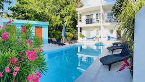Un área de piscina con sillones de descanso, una hamaca y un edificio de dos pisos con balcones.