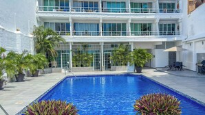 A swimming pool with a clear blue water, surrounded by potted plants, and a multi-story building with balconies in the background.