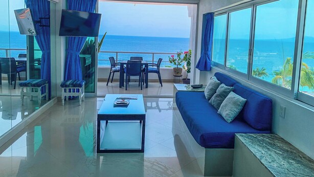 A modern living room with a sofa, coffee table, and TV, overlooking a balcony with a view of the sea.