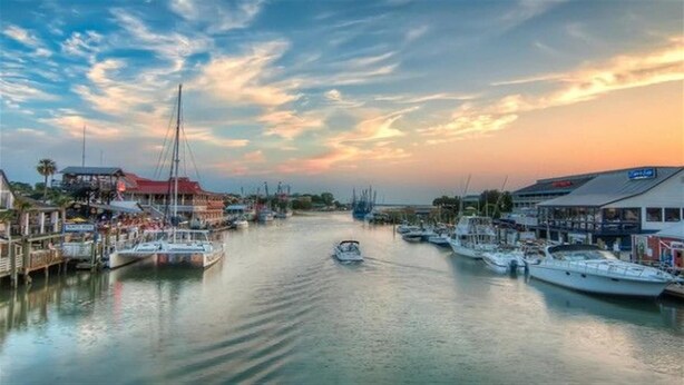 Una marina con barcos amarrados, un velero en el agua y edificios en la orilla.