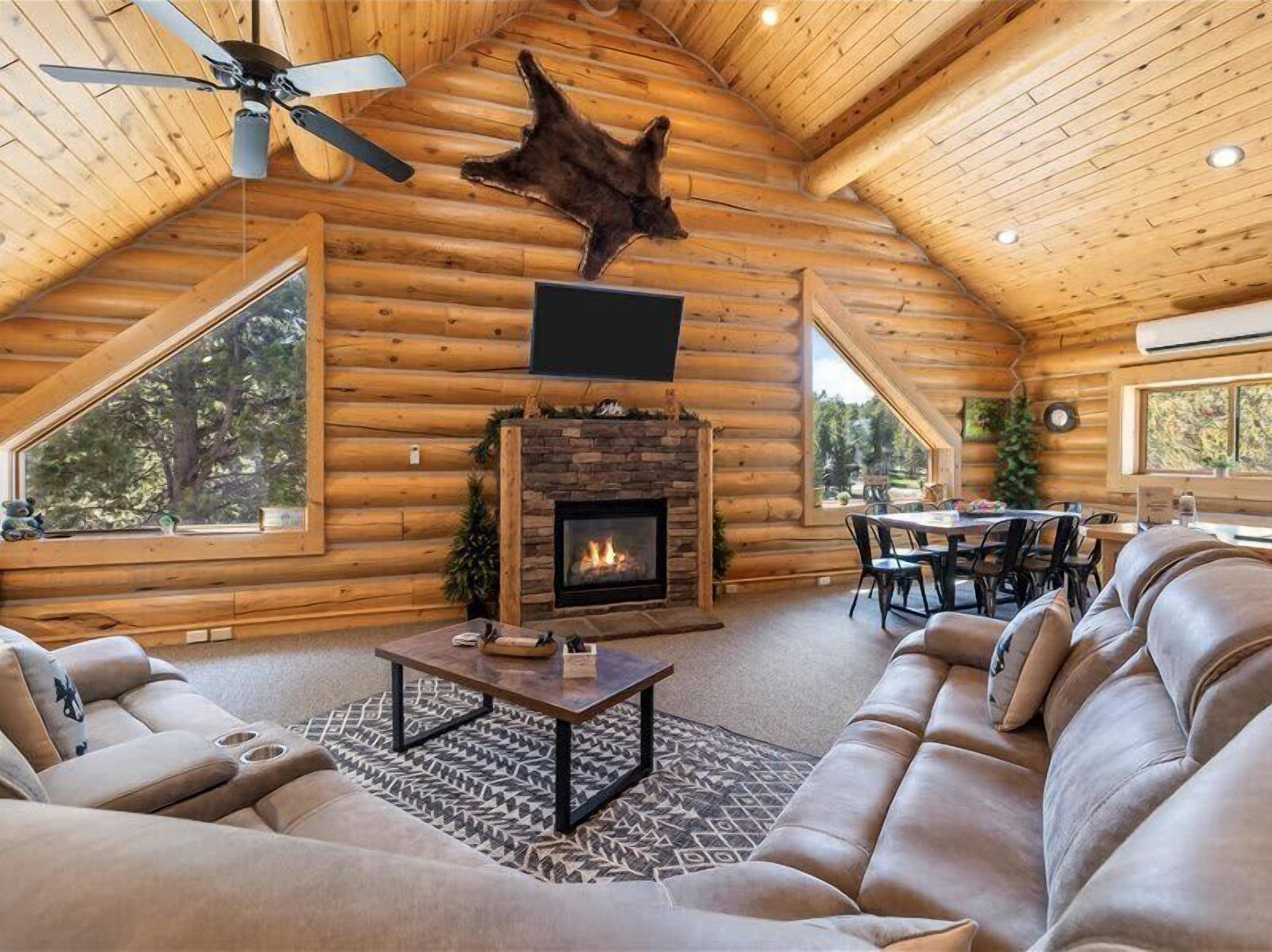 A log cabin interior with a fireplace, leather sofa, dining table, and large windows.