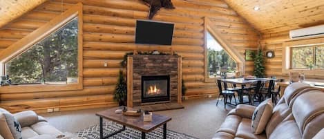Interior de una cabaña de troncos con chimenea, sofá de cuero, mesa de comedor y ventanas amplias.
