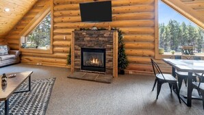 A log cabin interior with a stone fireplace, a mounted television, a sofa, a dining table with chairs, and a large window offering a forest view. - Bear Tracks - 20 Miles to Yellowstone - Air Condition - Wifi (Island Park)
