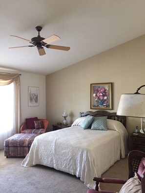 A bedroom with a bed, a nightstand, a chair, a ceiling fan, and a painting on the wall.