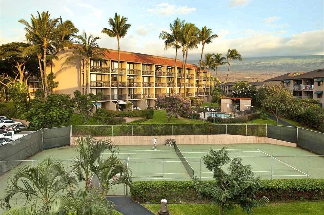 A hotel with multiple floors, a tennis court, and palm trees.