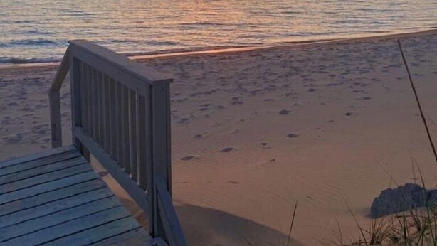 Ein hölzerner Steg führt zu einem Sandstrand, im Hintergrund ein Sonnenuntergang.
