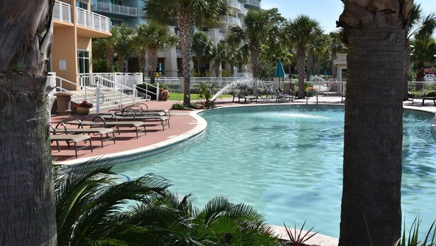 A swimming pool with a curved edge, surrounded by palm trees and a multi-story building with balconies. - Top Floor Legacy Penthouse / Stunning Gulf Views / Walk to Beach, Pools & more! (Gulfport)