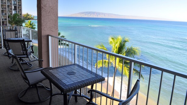 Un balcon avec une table et des chaises, donnant sur une plage bordée de palmiers et surmontée, au loin, d’une montagne.