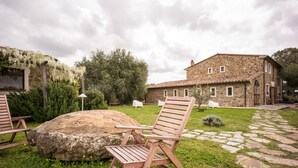 A stone building with a tiled roof, a garden with a large rock, and wooden chairs. - Tuscan Stone Farmhouse for 16 guests, amazing view, vineyards and olive trees (Manciano)