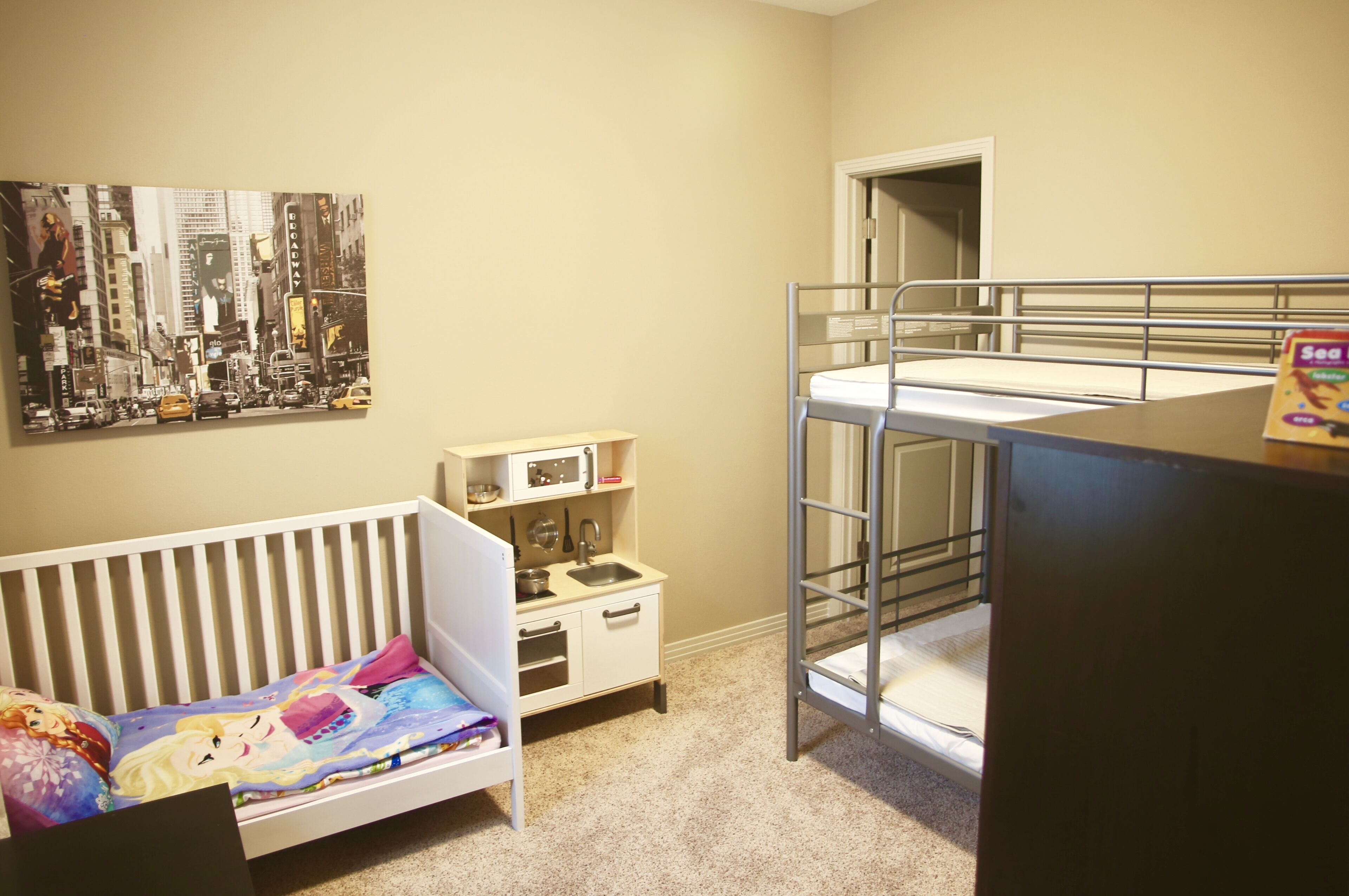 Een kinderkamer met een stapelbed, een ledikant, een kleine kitchenette en een stadsgezichtschilderij aan de muur.