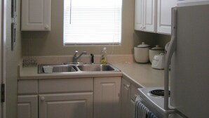 Una cocina compacta con armarios blancos, un lavamanos doble, un refrigerador y una ventana con persianas.