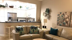A living room with a beige sofa, a coffee table with decorative items, a patterned rug, and a view of a modern kitchen with white cabinets and a stainless steel refrigerator. - Naples Vanderbilt Beach-beautiful Updated-walk to the beach (Naples)