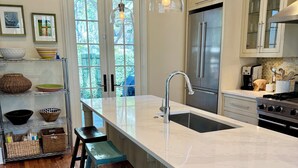 A kitchen with a large island, wooden stools, a stainless steel refrigerator, and hanging pendant lights.