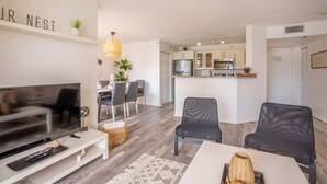 A modern living room with a flat-screen TV, a dining area with chairs, a kitchen with cabinets and appliances, and a hallway leading to a door.
