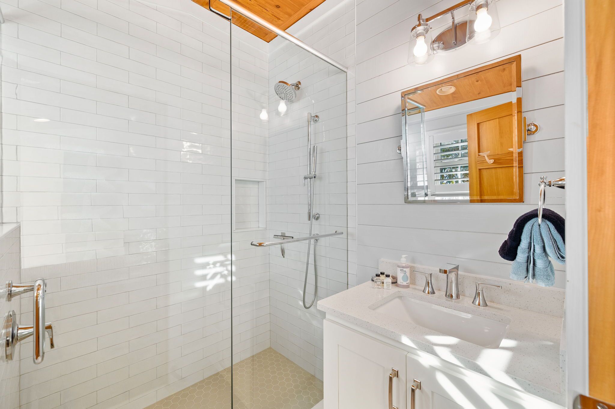 A modern bathroom with a glass-enclosed shower, white tiled walls, a wooden ceiling, and a vanity with a sink and mirror.