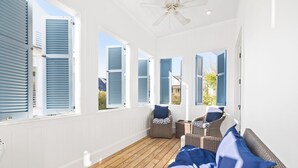 Ein sonnendurchflutetes Zimmer mit Holzboden, Korbmöbeln und großen Fenstern mit Rollläden.