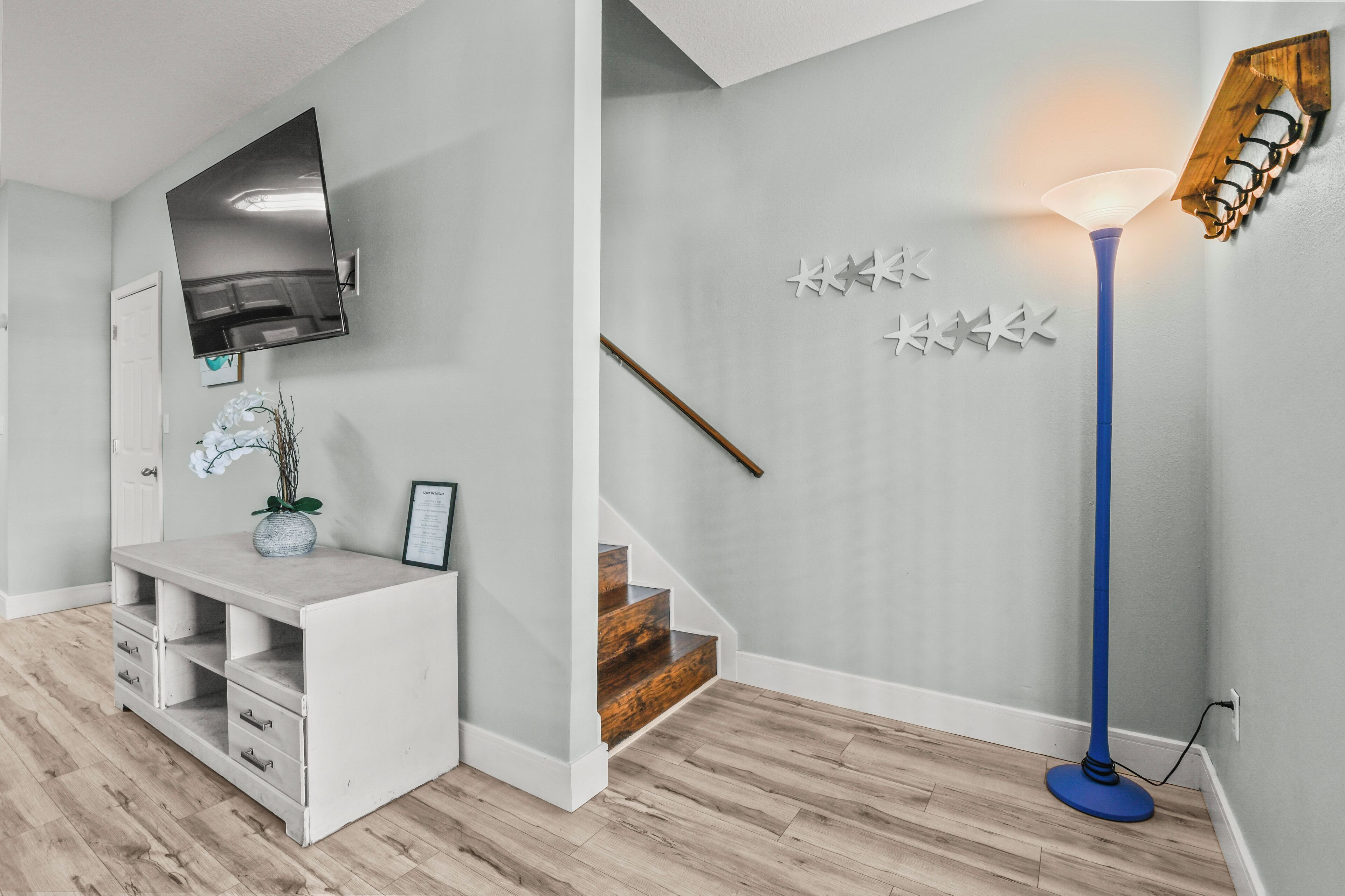 Un intérieur moderne avec un téléviseur à écran plat fixé au-dessus d’une console blanche, un lampadaire bleu et un escalier avec des marches en bois.