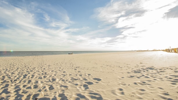 Uma praia arenosa com pegadas que levam à água, céu claro e um barco ao longe.