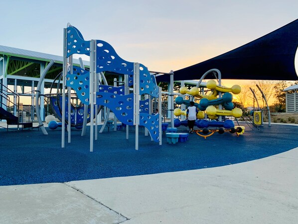 Un parque infantil con estructuras para escalar y toboganes, rodeado de una zona cubierta y un edificio al fondo.
