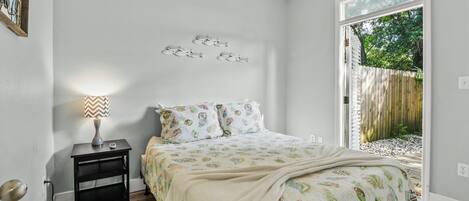 Et soveværelse med seng, et natbord, en lampe og et vindue med udsigt til et træhegn og grøntområde.