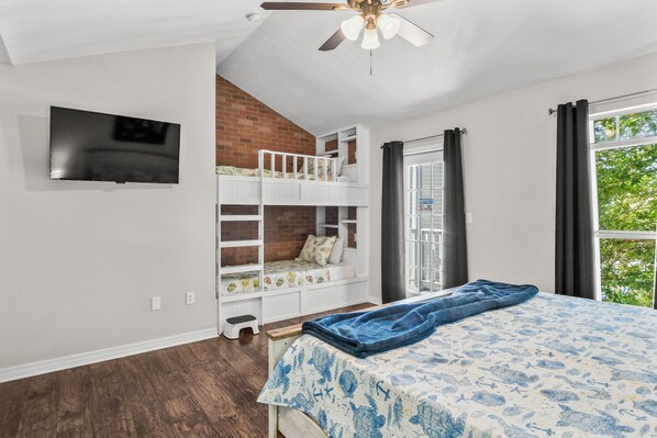 A bedroom with a bunk bed, a flat-screen TV mounted on the wall, a ceiling fan, and a window with curtains.