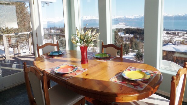 A round wooden dining table with chairs, a red vase with flowers, and plates on placemats. Large windows offer a view of a snowy landscape and mountains. - Miracle Ridge - Beautiful, Private Home Overlooking Kachemak Bay & Cook Inlet (Homer)