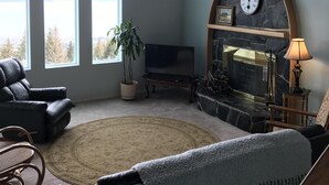 A living room with a fireplace, a television, and a large window with a view. - Miracle Ridge - Beautiful, Private Home Overlooking Kachemak Bay & Cook Inlet (Homer)