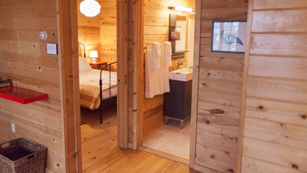 A wooden cabin interior with a bed, a small table with a lamp, a sink, and a window. - Fantastic House In Grand Marais! Walking Distance To Downtown! (Grand Marais)