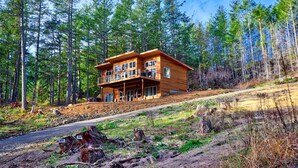 A two-story wooden cabin with a balcony, surrounded by a forest. - Water Views from this New Northwest Modern Home w/ Hot Tub (Eastsound)