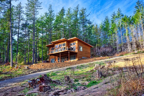 A two-story wooden cabin with a balcony, surrounded by a forest. - Water Views from this New Northwest Modern Home w/ Hot Tub (Eastsound)