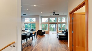 A spacious living area with a wooden floor, a kitchen with a bar, and large windows offering a view of trees. - Water Views from this New Northwest Modern Home w/ Hot Tub (Eastsound)