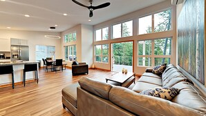 A spacious living area with a leather sofa, dining table, and chairs. There is a kitchen area with a refrigerator and a dining table. Large windows provide natural light and views of the outdoors. - Water Views from this New Northwest Modern Home w/ Hot Tub (Eastsound)