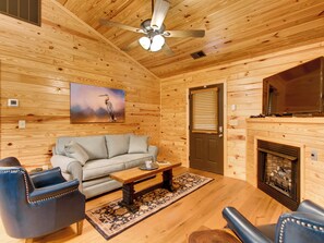 A cozy log cabin interior with a sofa, two armchairs, a coffee table, a fireplace, and a ceiling fan.