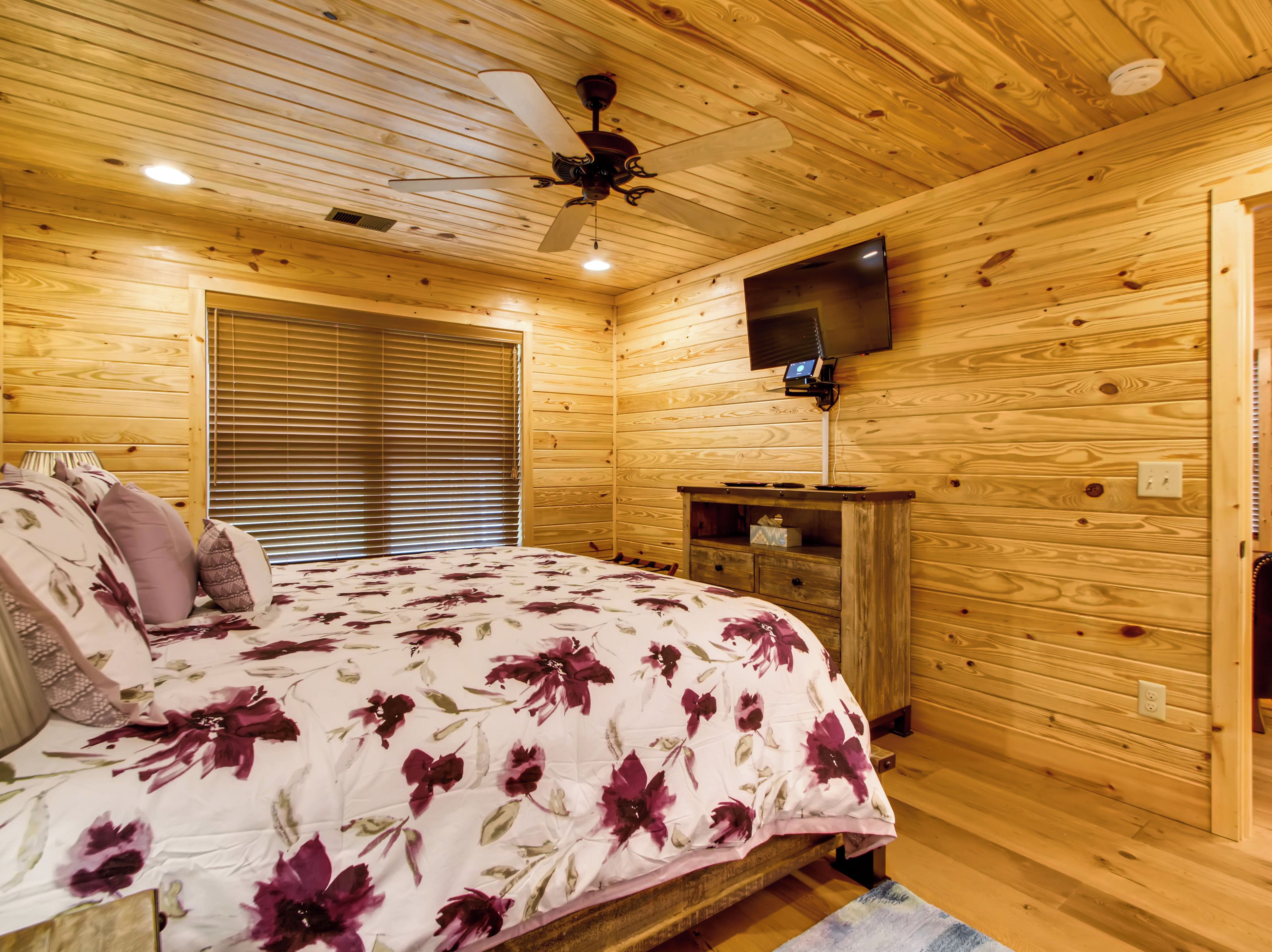 A wooden-paneled bedroom with a bed, a ceiling fan, a television mounted on the wall, and a wooden nightstand.