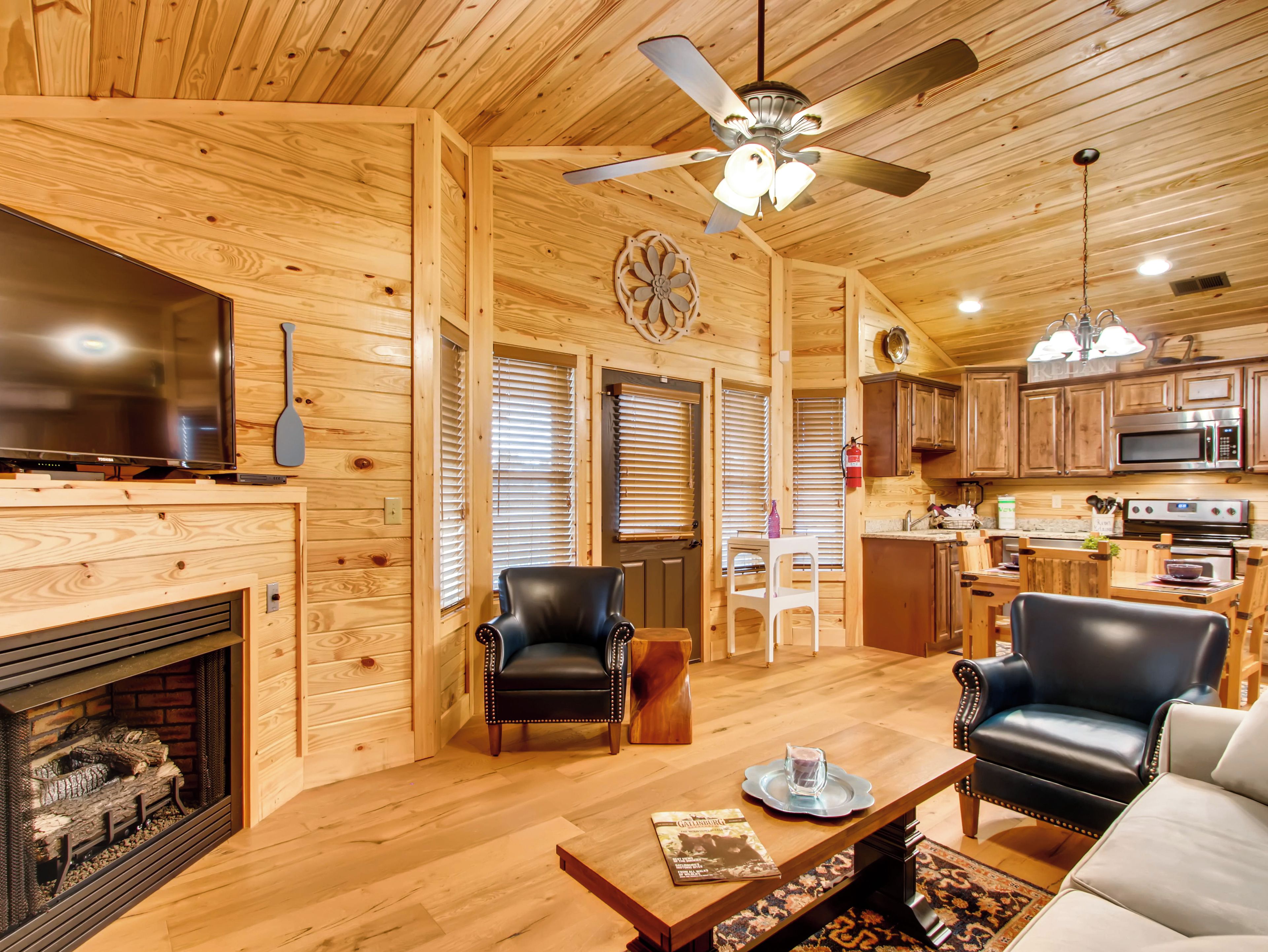 A cozy log cabin interior with a fireplace, leather chairs, a dining table, a ceiling fan, and wooden cabinetry.