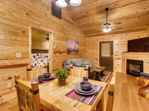 A cozy wooden cabin interior with a dining table set for two, a sofa, and a fireplace.