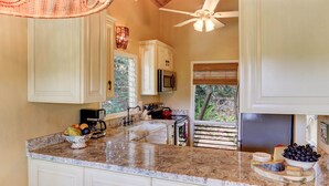 A kitchen with white cabinets, a granite countertop, a ceiling fan, and a window with a view of greenery. - Poipu Crater 26 | Tropical Retreat w/ Pool, Lanai & Garden Views (Koloa)