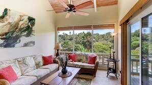 A spacious living room with a high ceiling fan, large windows offering a view of lush greenery, a sofa with red cushions, and a small table with a vase of flowers. - Poipu Crater 26 | Tropical Retreat w/ Pool, Lanai & Garden Views (Koloa)