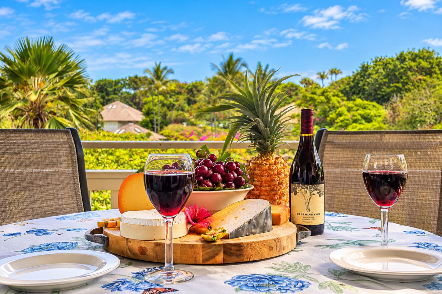 A table set with a bottle of wine, glasses, cheese, fruits, and a pineapple on a tablecloth with a floral pattern.