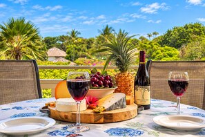 Une table dressée avec une bouteille de vin, des verres, du fromage, des fruits et un ananas, le tout posé sur une nappe à motifs floraux.