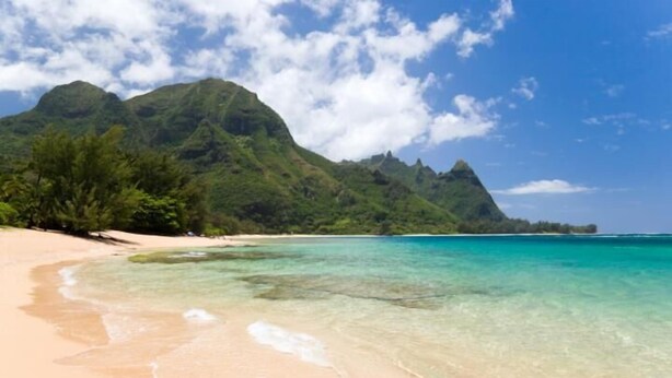 A beach with clear turquoise water, sandy shore, and lush green mountains in the background. - Poipu Crater 26 | Tropical Retreat w/ Pool, Lanai & Garden Views (Koloa)