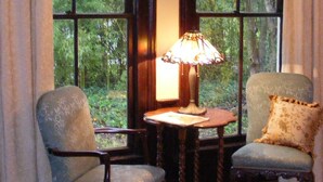Ein Zimmer mit zwei Stühlen, einem Tisch und einer Lampe. Große Fenster mit Vorhängen sind zu sehen.