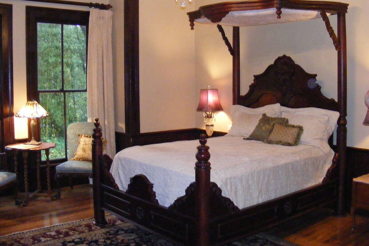A four-poster bed with a canopy, a nightstand with a lamp, a chair, and a view of greenery through the window.